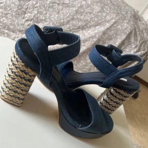 Blå sandaler med flätad klack från Axel - Snygga sandaler i blått denimtyg med breda remmar och justerbart spänne runt vristen. Klacken är hög och flätad i naturmaterial med blå detaljer för en trendig look. Perfekta för sommaren och ger en cool touch till outfiten. 