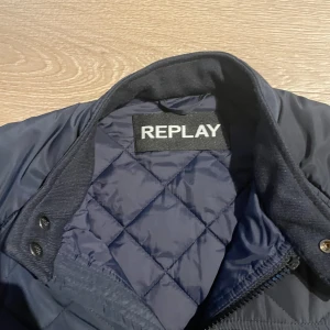 Mörkblå quiltad jacka från Replay - Snygg mörkblå quiltad jacka från Replay med dragkedja framtill och flera fickor med dragkedja. Jackan har hög krage med knappdetalj och ett stilrent rutmönster över hela framsidan.