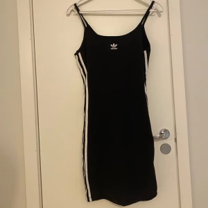 Svart klänning med smala axelband från Adidas - Svart tight klänning från Adidas med smala axelband och vita ränder längs sidorna. Liten Adidas-logga framtill. Sportig och stilren design som passar perfekt till sneakers.