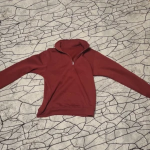 Vinröd halvzip tröja - Vinröd långärmad tröja med halv dragkedja i halsen. Tröjan har en hög krage och är i ett mjukt material, perfekt för kyligare dagar. Enkel och stilren design utan synliga loggor eller mönster.