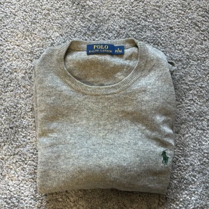 Grå tröja från Polo Ralph Lauren - Säljer en klassisk grå tröja från Polo Ralph Lauren med rund halsringning och diskret broderad logga i grönt på bröstet. Perfekt för en stilren och enkel look.