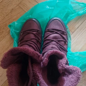 Vinröda boots med foder och snörning - Säljer ett par vinröda boots med mjukt foder och snörning framtill. Skorna har en rund tå och platt sula, perfekta för kyligare dagar. Utsidan är i mocka och insidan är fodrad för extra värme.