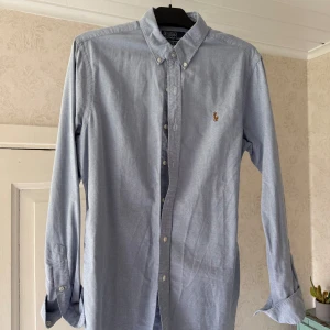 Ljusblå skjorta från Polo Ralph Lauren - Klassisk ljusblå skjorta från Polo Ralph Lauren med button-down krage och broderad logga på bröstet. Slim fit-modell med långa ärmar och vita knappar. Perfekt för en stilren look.
