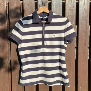 Randig pikétröja från Tommy Hilfiger - Säljer en klassisk pikétröja från Tommy Hilfiger i marinblått och vitt med breda horisontella ränder. Tröjan har korta ärmar, dragkedja framtill och liten logga på bröstet. 