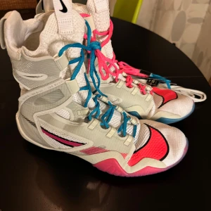 Nike hyper ko 2 LE  - Säljer ett par Nike boxing skor i vitt med rosa och blå detaljer. Skorna har snörning med både blå och rosa skosnören, samt svarta Nike-loggor. Ovandelen är i syntetmaterial med meshpaneler för extra ventilation. Perfekta för dig som vill sticka ut på ringen! Priset kan diskuteras 