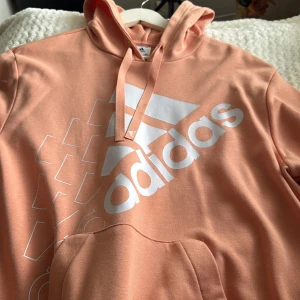 Persikofärgad hoodie från Adidas - Säljer en snygg persikofärgad hoodie från Adidas med stor vit logga framtill och en stor magficka. Perfekt för en avslappnad stil.