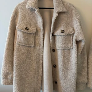 Beige teddyjacka med knappar - Säljer en beige teddyjacka med stora bröstfickor och knappar framtill. Jackan har krage och lång ärm, perfekt för kyligare dagar. Mysig och trendig modell som passar till många outfits.