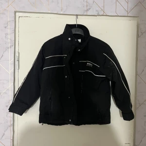 Svart vinterjacka från H&M med reflexdetaljer - Svart vinterjacka från H&M med hög krage och dragkedja samt knappar framtill. Jackan har vita reflexdetaljer på bröstet och ryggen samt två fickor med dragkedja. Perfekt för kalla dagar.