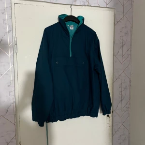 Mörkblå vindjacka från Skiing - Retro vindjacka i mörkblått med turkosa detaljer och halv dragkedja framtill. Jackan har hög krage, lång ärm och en stor ficka med knappar på magen. Perfekt för kyliga dagar och enkel att dra över huvudet.