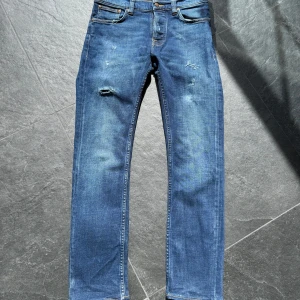 Nudie Grim Tim  - Säljer ett par klassiska blå jeans med raka ben och lätt slitna detaljer framtill. Jeansen har fem fickor, normal passform och stängs med dragkedja och knapp. Perfekta för en avslappnad stil.