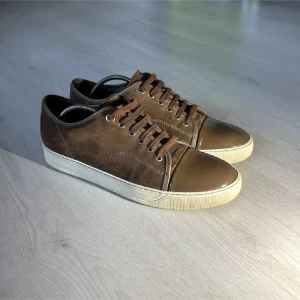 Bruna Lanvins - Säljer ett par bruna sneakers från Lanvin med ovandel i mocka och tåparti i blankt skinn. Skorna har snörning och en vit platt sula. Perfekta för dig som gillar stilrena och klassiska sneakers med en twist. Skriv vid minsta lilla fundering