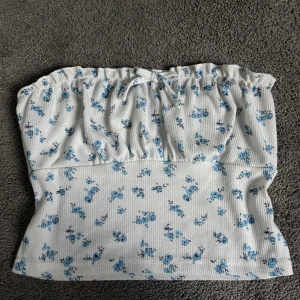 Vit blommig tubtopp med blå blommor - Supersöt vit tubtopp med små blå blommor och rynkdetalj upptill. Toppen har en liten knytning framtill och är gjord i ett ribbat material som ger en skön passform. Perfekt för varma dagar och somriga outfits!