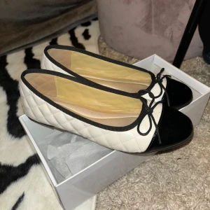 Svartvita ballerinaskor med rosett - Säljer ett par helt nästintill nya klassiska ballerinaskor i quiltad aprikos syntet med svarta detaljer och lackad tå. Skorna har en söt rosett framtill och rund tå. Perfekta för en stilren look. 