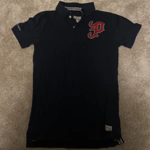 Svart pikétröja från Peak Performance - Klassisk svart pikétröja från Peak Performance med röd broderad P-logga på bröstet och krage med knappar.  Använd fåtal gånger.