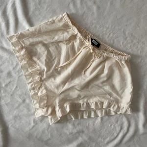 Beige volangshorts med snörning från BIKBOK - Säljer ett par ljusbeige shorts från BIKBOK med volangdetaljer längs kanterna och snörning i midjan. De är tillverkade i ett tunt och mjukt tyg, perfekta för varma dagar.