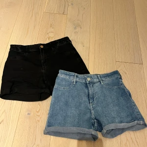 Två par jeansshorts i svart och blå denim - Säljer två par klassiska jeansshorts, ett par i svart denim och ett par i ljusblå denim. Båda har uppvikta benslut, femficksmodell, knappgylf och är hightwaist. Perfekta för sommaren och enkla att matcha med olika toppar!🤩