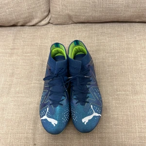 Blåa fotbollsskor från Puma - Säljer ett par blåa fotbollsskor från Puma med vita detaljer och gröna insidor. Skorna har snörning, rund tå och platt sula med dobbar för bra grepp på planen. Perfekta för dig som vill sticka ut på matchen!
