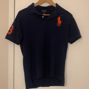 Mörkblå pikétröja från Polo Ralph Lauren - Säljer en mörkblå piké från Polo Ralph Lauren med orange logga på bröstet och en orange siffra 3 på högra ärmen. Klassisk krage och två knappar vid halsen. Perfekt för en avslappnad och stilren look. Storleken är L (14-16 år i barnstorlek) men jag skulle säga att det sitter ungefär som XS. Jag uppskattar att pikén passar på dig som är runt 165-170cm och väger runt 55kg. Skicket skulle jag säga är ungefär 7,5/10