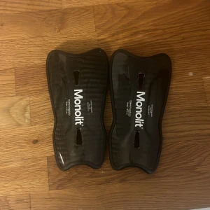 Svarta benskydd från Monolit - Säljer ett par svarta benskydd från Monolit. De har en ergonomisk form och är tillverkade i syntetmaterial. Perfekta för fotbollsträning eller match. Snygg och stilren design med vit logga på framsidan.