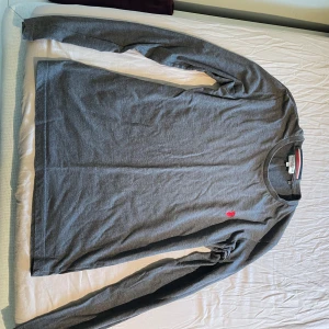 Grå långärmad tröja från U.S. Polo Assn - Säljer en stilren grå långärmad tröja från U.S. Polo Assn med en liten röd logga broderad på bröstet. Tröjan har rund halsringning och är perfekt för en enkel och klassisk look.