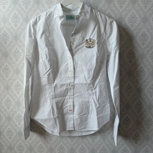 Vit skjorta från Morris - Snygg vit skjorta från Morris med broderad detalj på bröstet. Skjortan har en klassisk krage och knappar framtill. Perfekt för en stilren look. Rökfritt hem! Använd fåtal gånger!
