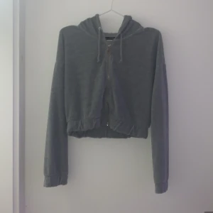 Grå croppad hoodie från Bershka - Säljer en stilren grå croppad hoodie från Bershka. Den har en dragkedja framtill och en justerbar huva med snören. Perfekt för en avslappnad look! Liten skavank på ena sidan (se sista bilden) men inget som påverkar passformen eller syns på långthåll.