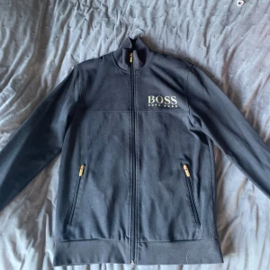 Svart kofta från Hugo Boss - Säljer en stilren svart kofta från Hugo Boss med dragkedja och två fickor med gulddetaljer. Koftan har en hög krage och långa ärmar, perfekt för en casual look. Märkesloggan är broderad på bröstet. SKRIV OM NI UNDRAR NÅGOT MER 