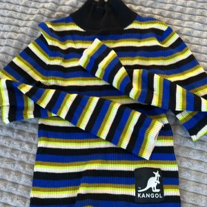 Randig ribbad tröja från Kangol - Säljer en färgglad randig tröja från Kangol med ribbad struktur. Tröjan har en hög krage och långa ärmar. Färgerna är blå, gul, svart och vit. Perfekt för att liva upp garderoben!
