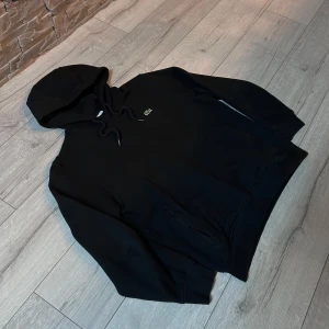 Svart Lacoste hoodie  - Säljer en stilren svart hoodie från Lacoste med det klassiska krokodilmärket på bröstet. Tröjan har en justerbar huva med snören och ribbade muddar vid ärmslut och nederkant. Perfekt för en avslappnad look. 
