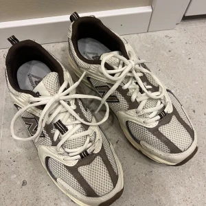 Vita och bruna sneakers från New Balance - Säljer ett par stilrena New Balance sneakers i vitt och brunt. Skorna har en klassisk design med snörning och meshdetaljer. Använda 2 gånger så de är som nya, kartong finns kvar och kommer skickas i den! Säljes pga ångrat köp. Nypris 1399.