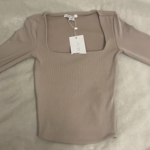 Beige ribbad topp från Amisu - Säljer en stilren beige ribbad topp från Amisu. Toppen har en fyrkantig urringning och långa ärmar, perfekt för en elegant look. Materialet är mjukt och stretchigt för en bekväm passform.