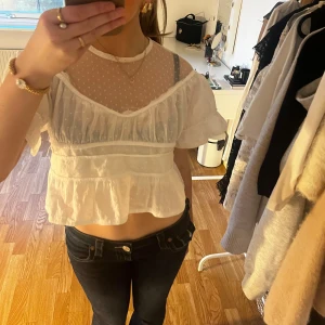 Vit blus med spetsdetaljer - Charmig vit blus med spetsdetaljer och prickigt mönster på övre delen. Blusen har korta ärmar och en volangkant nedtill som ger en romantisk touch. Perfekt för en somrig look.