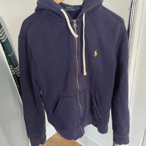 Mörkblå hoodie från Polo Ralph Lauren - Säljer en mörkblå hoodie från Polo Ralph Lauren med dragkedja och huva. Tröjan har en liten gul logotyp på bröstet och vita snören vid huvan. Perfekt för en avslappnad stil.