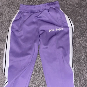 Lila byxor från Palm Angels - Snygga lila träningsbyxor från Palm Angels med vita och svarta ränder längs sidorna. Byxorna har en elastisk midja och sidofickor för extra bekvämlighet. Perfekta för en sportig look.