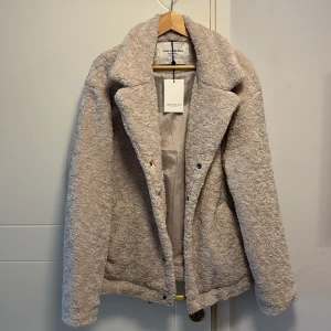 Beige teddyjacka från These Glory Days - Säljer en mysig beige teddyjacka från These Glory Days. Jackan har en mjuk och fluffig yta med långa ärmar och en klassisk krage. Perfekt för kyliga dagar när du vill hålla dig varm och stilren.