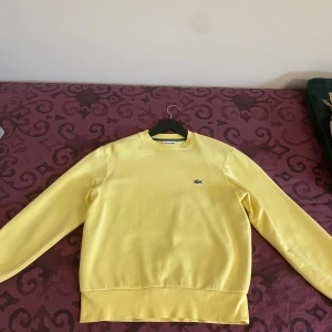 Gul tröja från Lacoste - Säljer en stilren gul tröja från Lacoste med klassisk passform. Tröjan har långa ärmar och en liten broderad logotyp på bröstet. Perfekt för en avslappnad stil. Storlek S