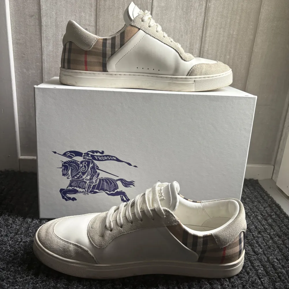 Snygga vita sneakers från Burberry med klassiskt rutigt mönster på hälen. Skorna har snörning och en stilren design med detaljer i mocka och syntetmaterial. Perfekta för en trendig look. Köpte för 6800 Allt Og ingår. Kengät.