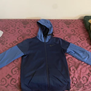 Blå träningsjacka från Nike - Säljer en blå träningsjacka från Nike med huva och dragkedja. Jackan har långa ärmar och är tillverkad i Dri-FIT-material för optimal komfort under träning. Perfekt för aktiva dagar! Storlek S men kan även passa M