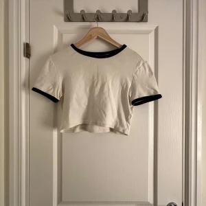 Vit croppad t-shirt från Zara - Säljer en vit croppad t-shirt från Zara med svarta kanter vid hals och ärmslut. Perfekt för en avslappnad stil. T-shirten har korta ärmar och en enkel design.