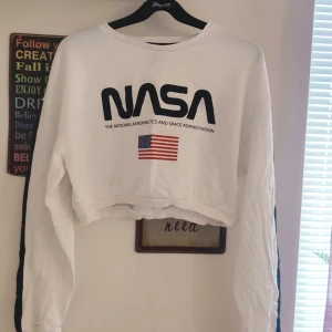 Vit croppad tröja med NASA-tryck - Säljer en vit croppad tröja från Divided med NASA-tryck och amerikansk flagga på framsidan. Tröjan har långa ärmar och en avslappnad passform. Perfekt för en cool och avslappnad stil. 🌟