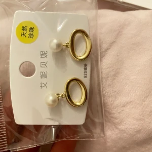 Guldörhängen med pärlor - Eleganta örhängen i guld med hängande vita pärlor. Perfekta för att ge en touch av lyx till din outfit. De har en oval form och är lätta att bära.