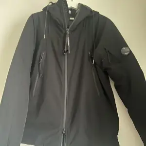 Snygg svart jacka från C.P. Company med dragkedja och huva. Tillverkad i högpresterande stretchpolyester med WR-beläggning och Primaloft-foder för extra värme. Perfekt för lite kyligare och vindigare dagar, tags finns 