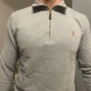 Grå tröja med dragkedja från Ralph Lauren - Säljer en stilren grå tröja från Ralph Lauren med dragkedja och broderad logga på bröstet. Tröjan har långa ärmar och en klassisk krage. Perfekt för en avslappnad men ändå elegant look.