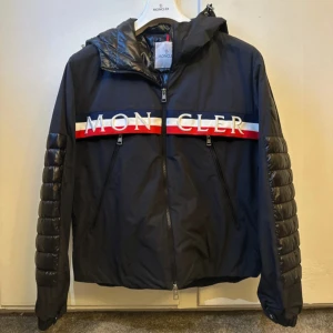 Svart jacka från Moncler - Snygg svart jacka från Moncler med dragkedja och huva. Jackan har långa ärmar med glansiga detaljer och Moncler-logga på bröstet. Den har röd, vit och blå rand över bröstet med Moncler-text. Storlek 2 motsvarar S