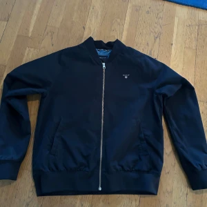 Marinblå bomberjacka från GANT storlek 158/164cm - Snygg marinblå bomberjacka från GANT med dragkedja framtill och broderad logga på bröstet. Jackan har en klassisk design med ribbade muddar och en stilren look. Perfekt för en casual stil.