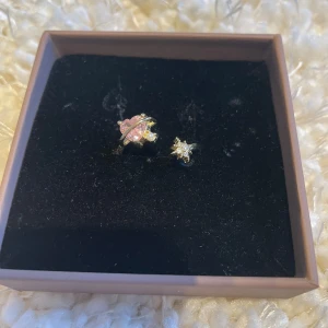Guldring med rosa och vita stenar - Unik guldring med en stor rosa sten och en mindre vit sten. Designen har en stjärnliknande form och en elegant guldfinish. Perfekt för att ge en touch av glamour till din stil.