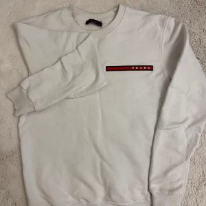 Vit tröja från Prada. Lite fläckar på men annars bra. - Säljer en stilren vit tröja från Prada med en röd och svart logga på bröstet. Tröjan har långa ärmar och ribbade muddar vid ärmslut och nederkant. Perfekt för en minimalistisk look.