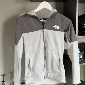 Grå och vit zip hoodie från The North Face - Säljer en snygg grå och vit zip hoodie från The North Face med huva och dragkedja. Hoodien  har en modern design med kontrasterande färger och är perfekt för kyligare dagar. Den har långa ärmar och är tillverkad i ett syntetmaterial.