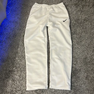 Vita träningsbyxor från Nike - Snygga vita träningsbyxor från Nike med elastisk midja och det klassiska Nike-logotypen på benet. Perfekta för sport och avslappning. Byxorna har en rak passform och är tillverkade i ett mjukt syntetmaterial.
