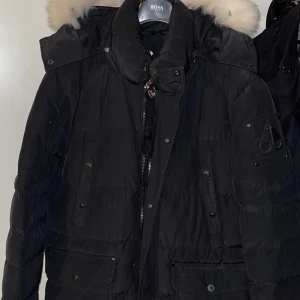 Svart dunjacka från Moose Knuckles - Säljer en svart dunjacka från Moose Knuckles med pälskantad huva. Jackan har dragkedja och knappar framtill samt flera praktiska fickor. Perfekt för kalla dagar!
